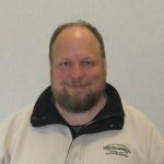 Male with brown hair and beard wearing a white coat. 